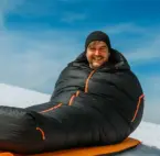 Ein Mann liegt in einem Schlafsack auf einer Isomatte in einer Schneelandschaft.